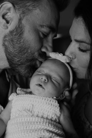 Monochrome image of parents kissing their sleeping newborn wrapped in knit fabric.