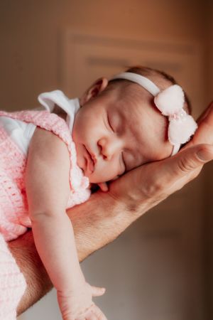 Peaceful sleeping newborn in pink dress and white bow headband resting on parent's arm.