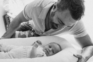 Black and white photo of parent leaning over and smiling at a baby on white bedding.