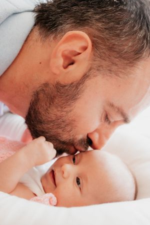 A tender moment between a bearded parent and infant lying on white bedding.