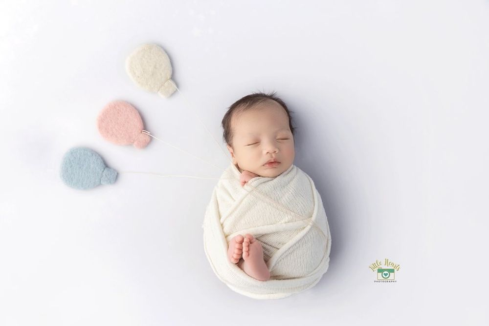 Sleeping newborn wrapped in white fabric with pastel balloons on white background.