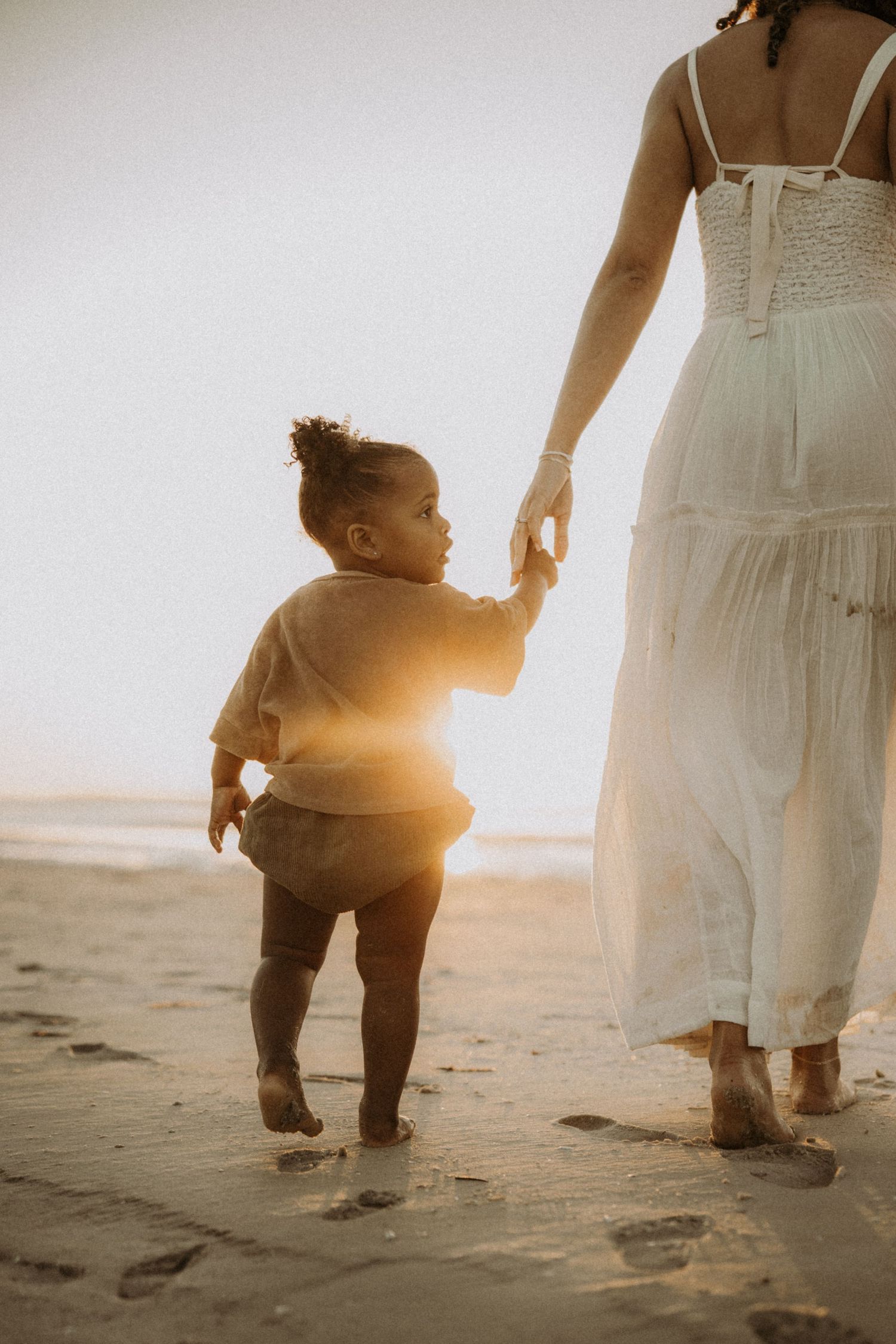 A parent and toddler walk along a sunlit beach at sunset leaving footprints in the sand.