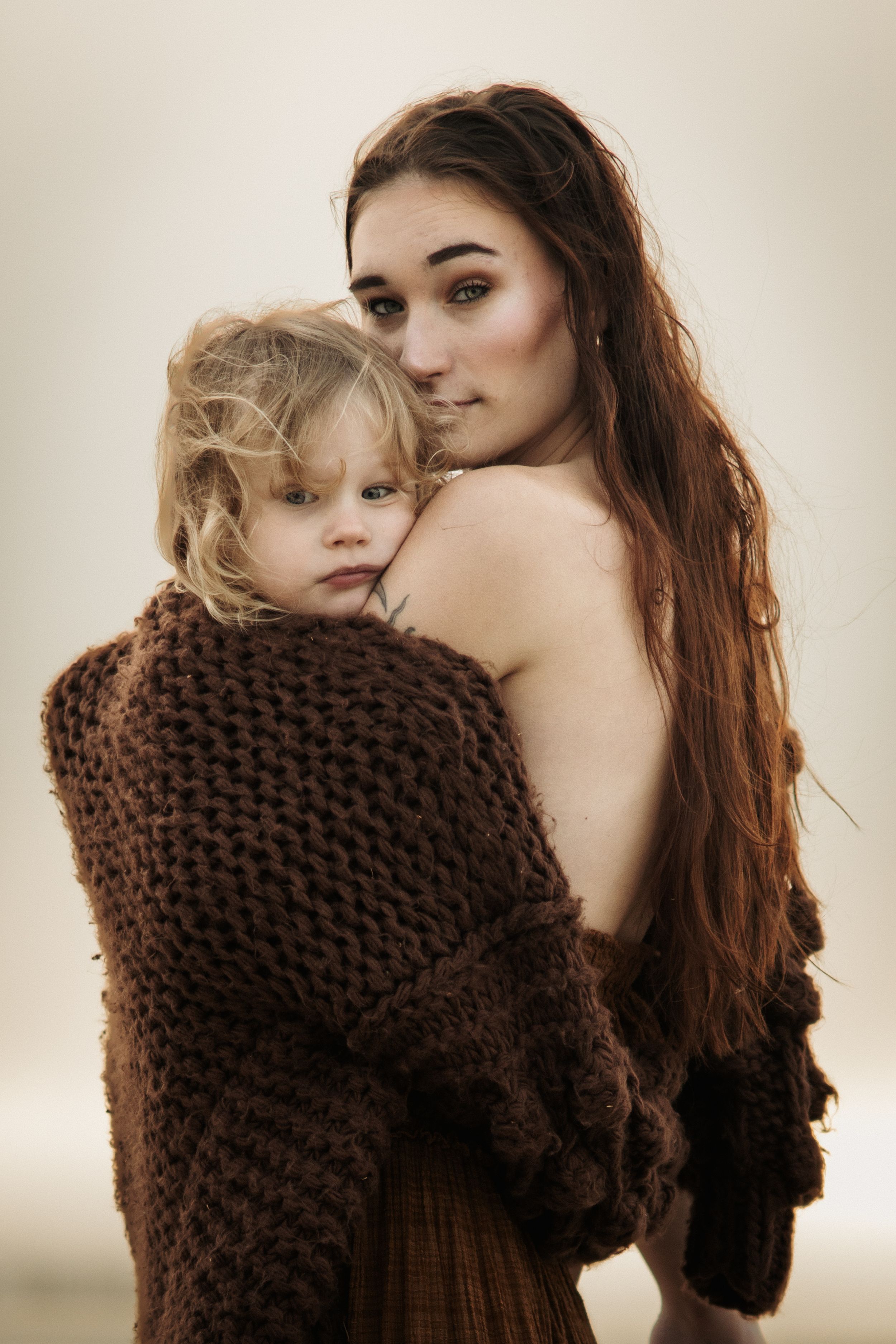A woman in a brown knit sweater embraces a small child against a soft beige background.