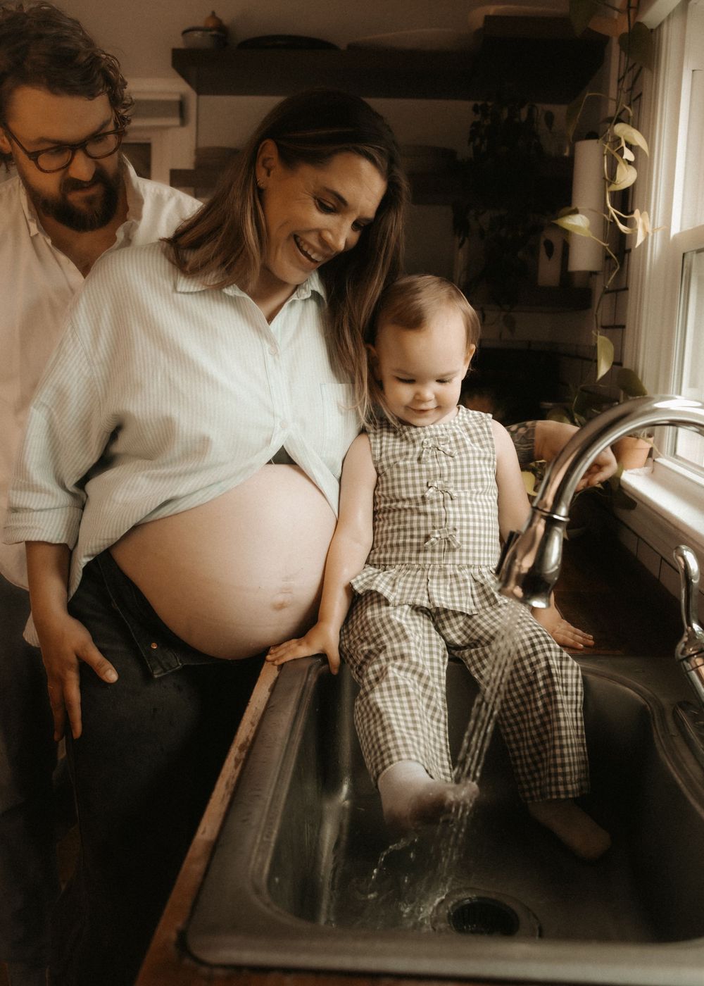 Pregnant woman and man smile at toddler sitting in kitchen sink with running water during family maternity photo session.