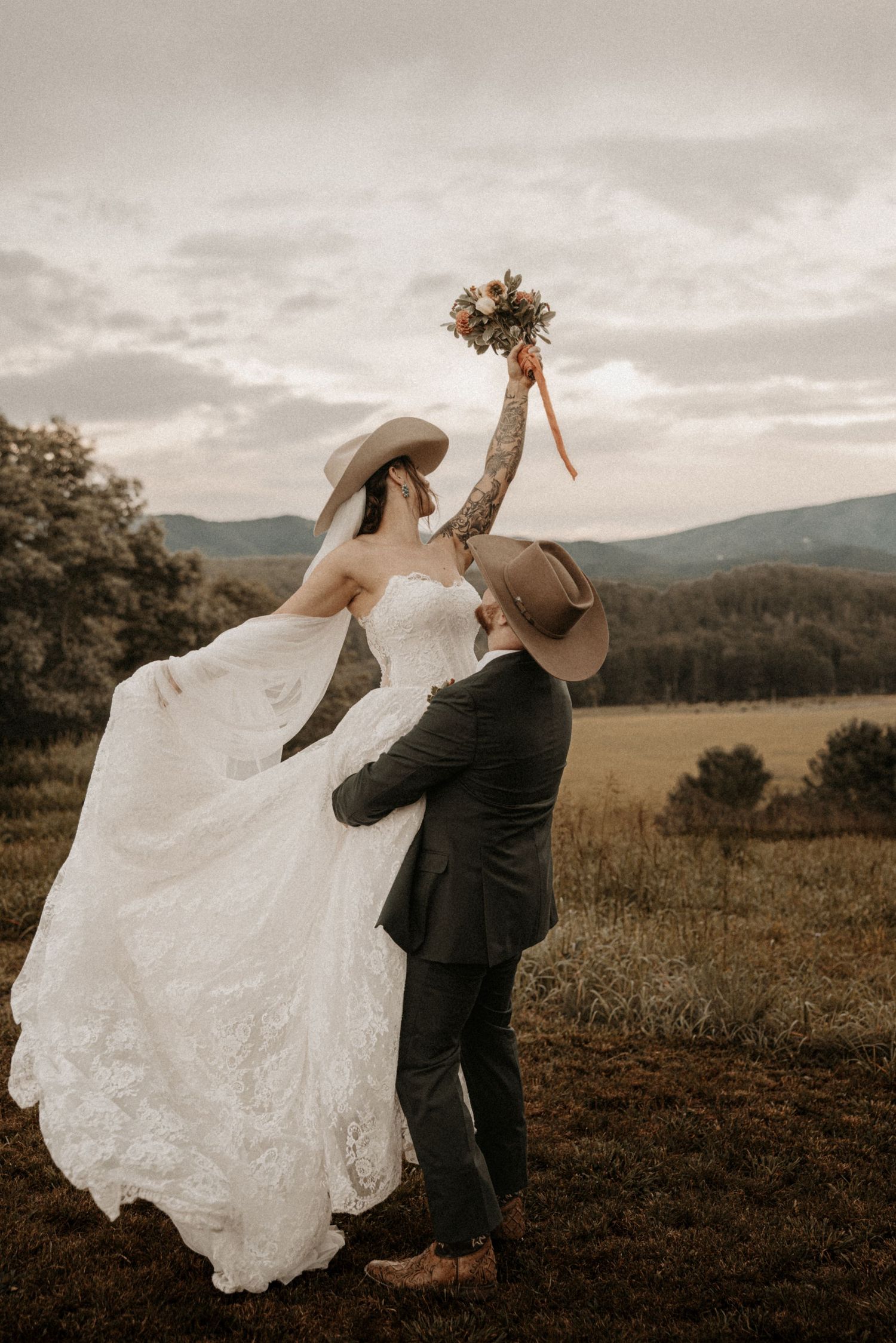 Bride and groom in cowboy hats celebrate outdoors, groom lifting bouquet with mountain backdrop.