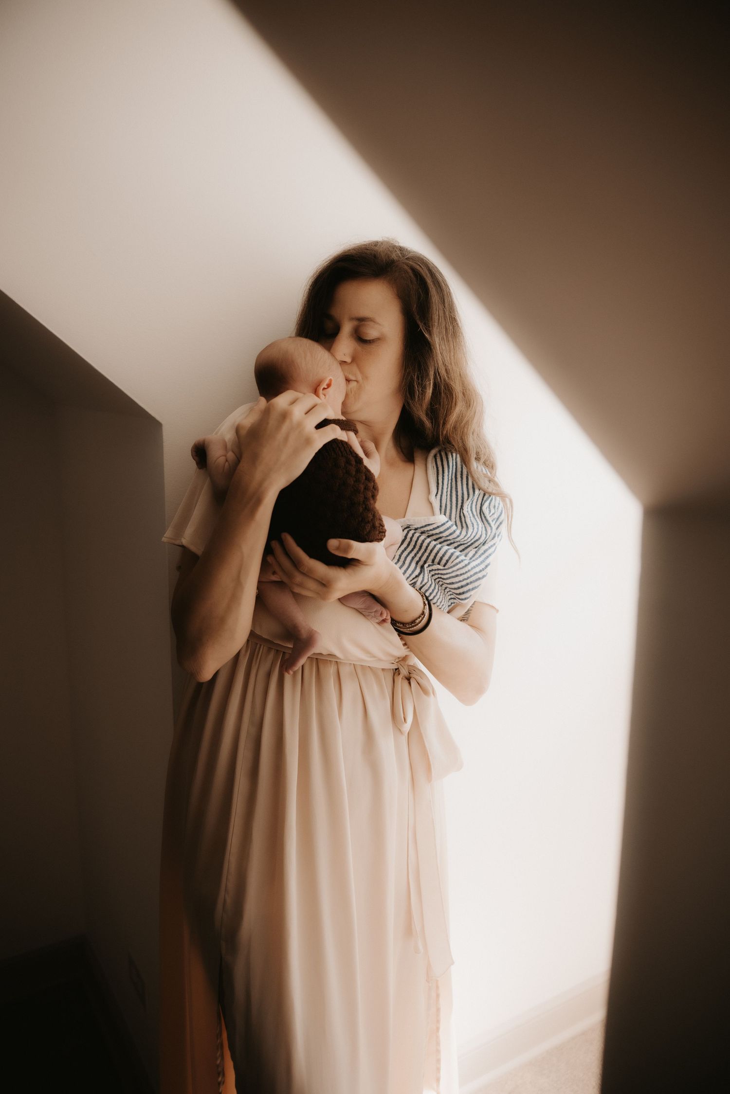 A mother tenderly kisses her newborn baby while standing in soft natural light wearing a blush pink skirt.