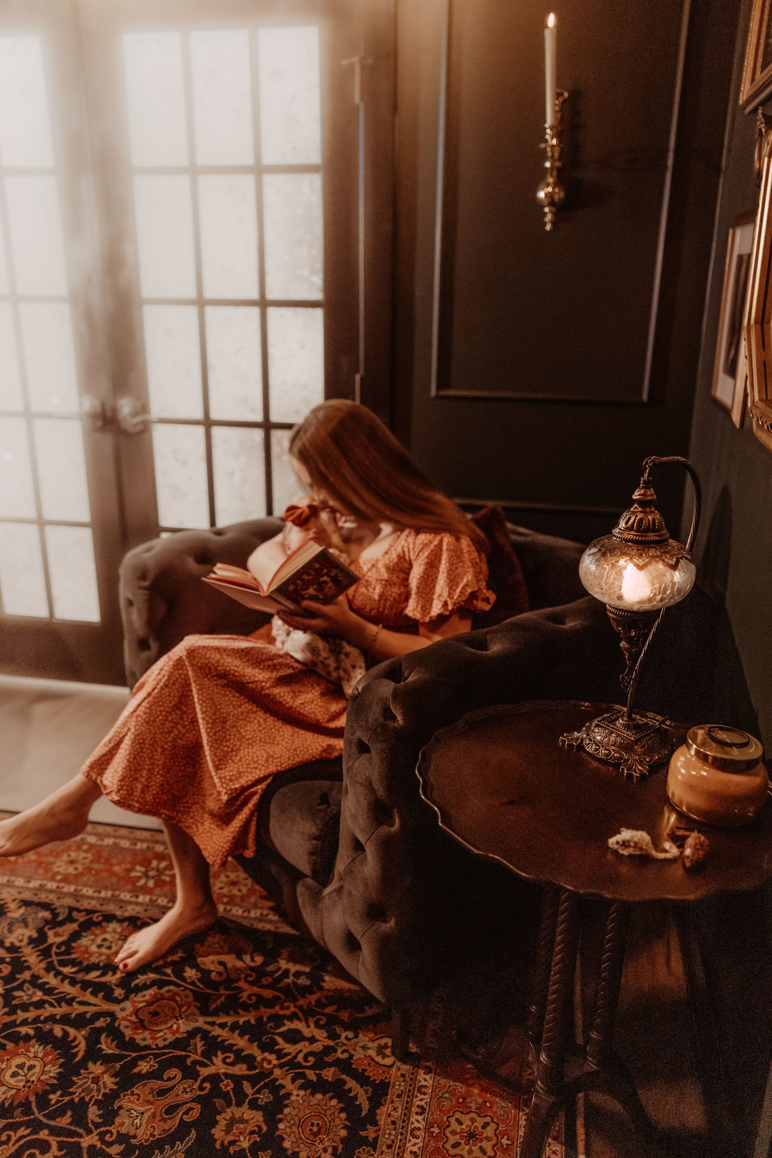 A black dog lies on a cozy brown couch near sunlit windows with a patterned rug below.