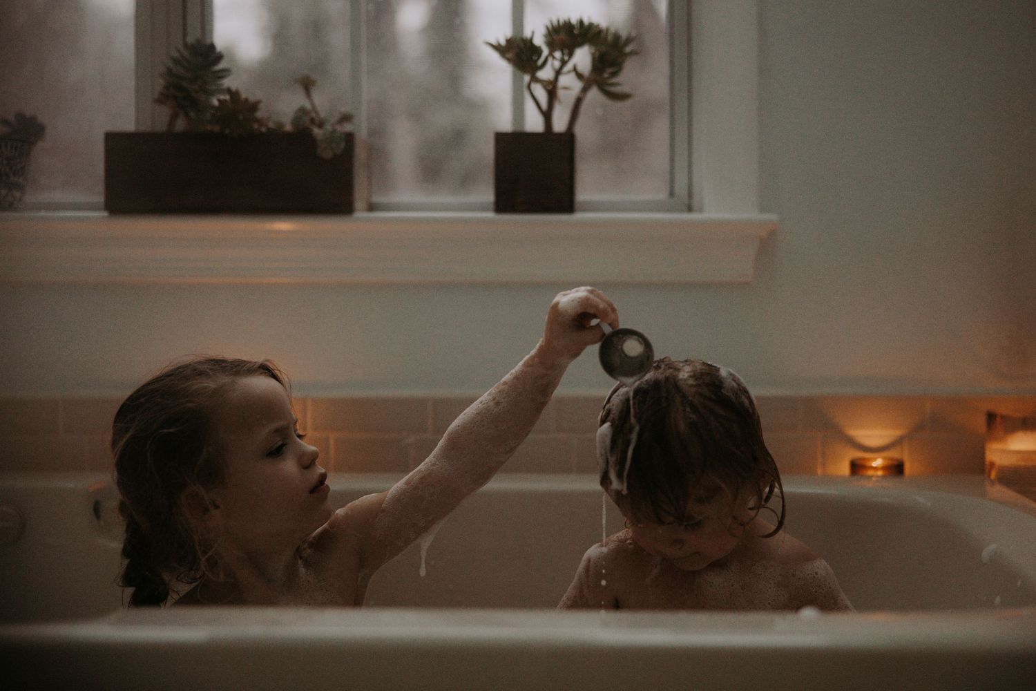 Two young children bathing together in a candlelit bathtub, one pouring water over the other's head.