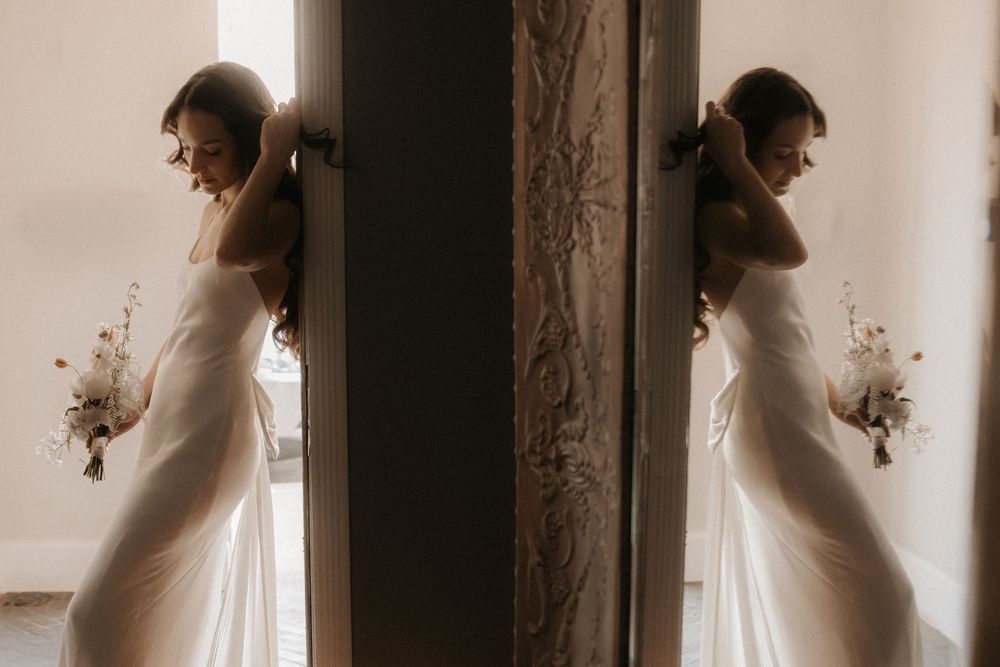 Bride in white gown reflected in mirror holding bouquet, elegant wedding portrait with natural light.