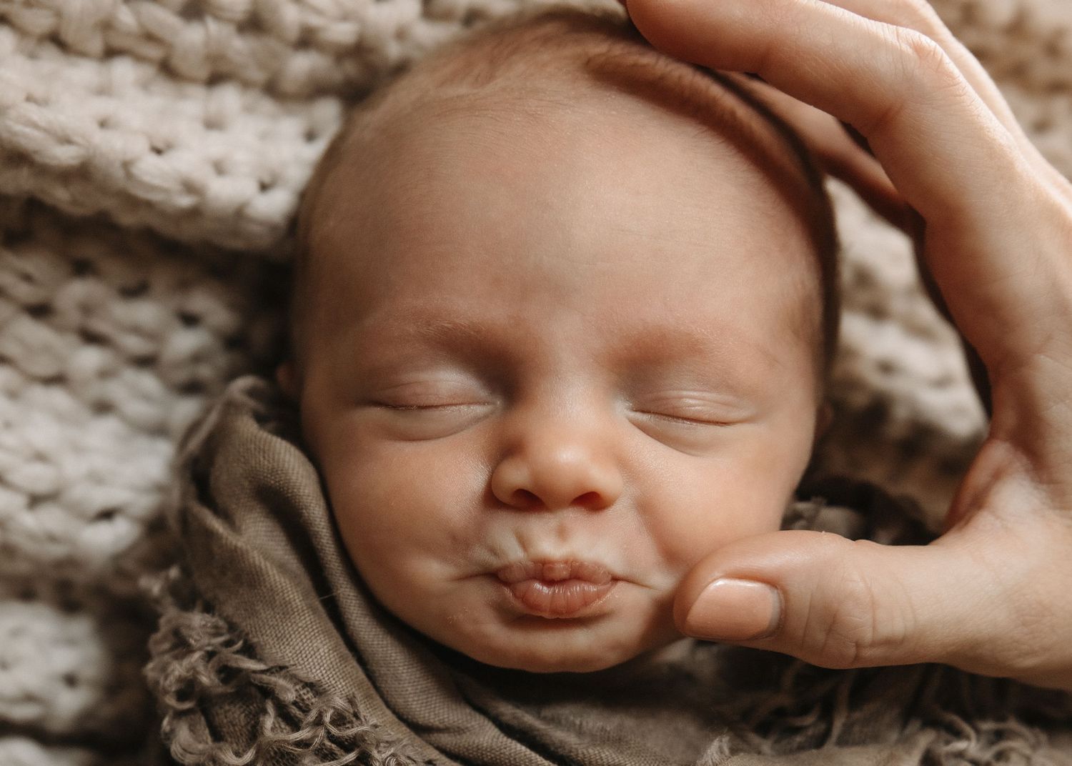 Peaceful newborn baby sleeping wrapped in soft taupe fabric with adult hand gently cradling head on knit blanket.