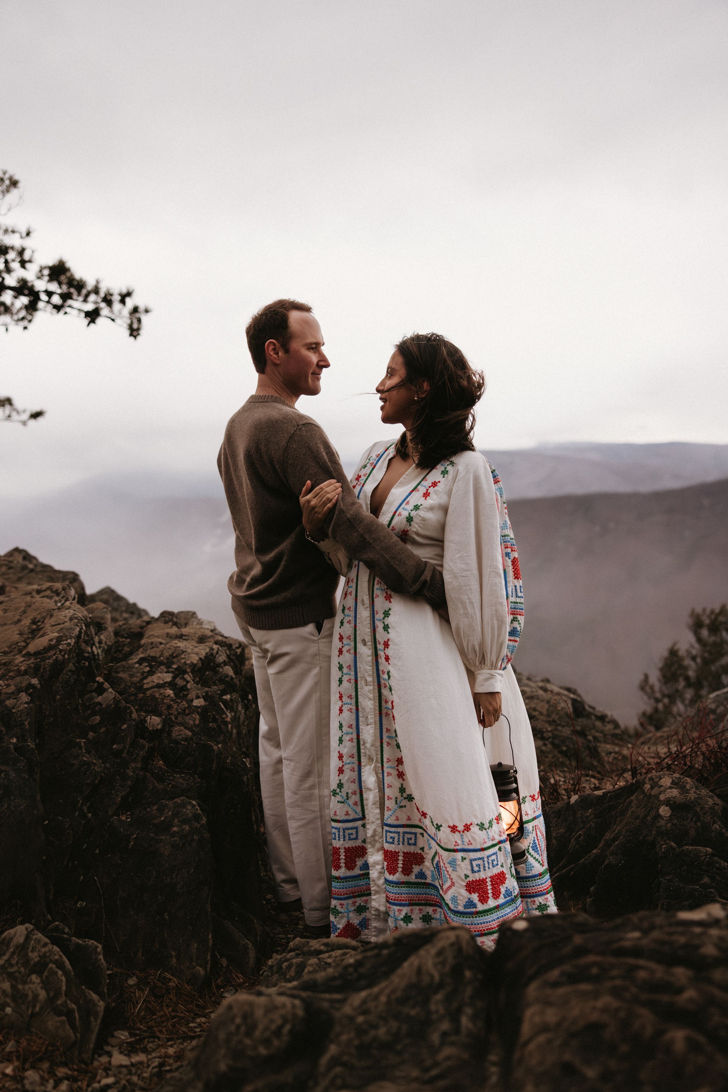 A couple in traditional embroidered clothing embraces against a mountainous landscape at sunset.