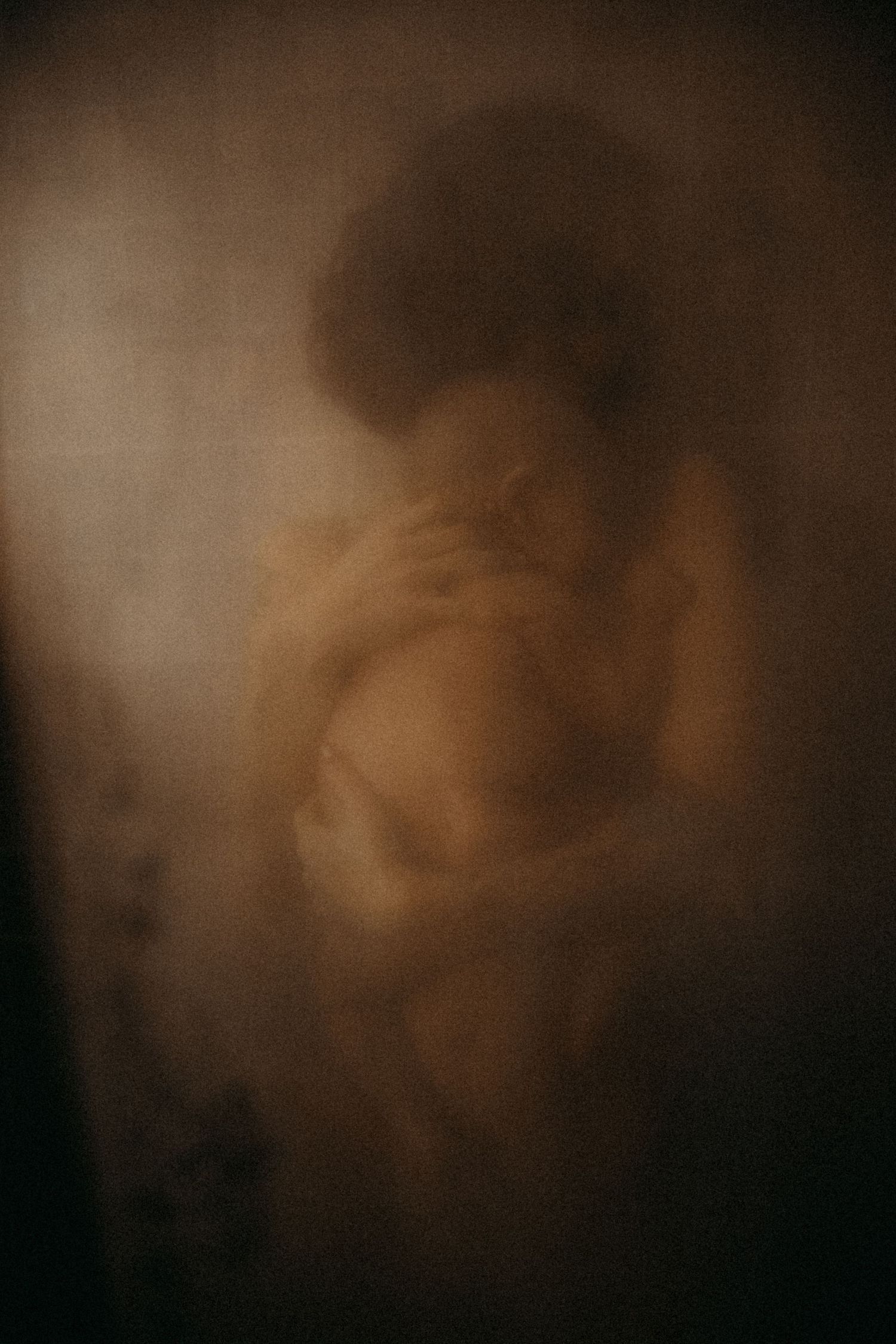 A blurred, ethereal silhouette of a person wrapped in fabric, bathed in warm sepia tones, creating a dreamy, mysterious mood.