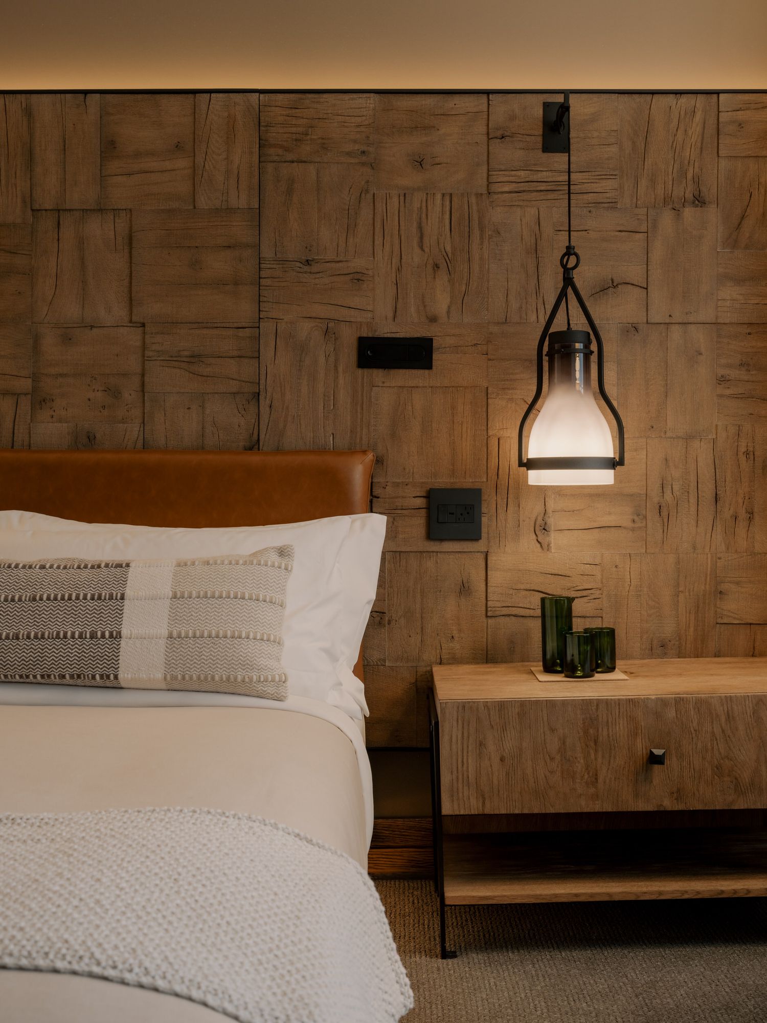 Modern bedroom with wooden paneled wall, white bedding, and hanging pendant light fixture beside wooden nightstand.