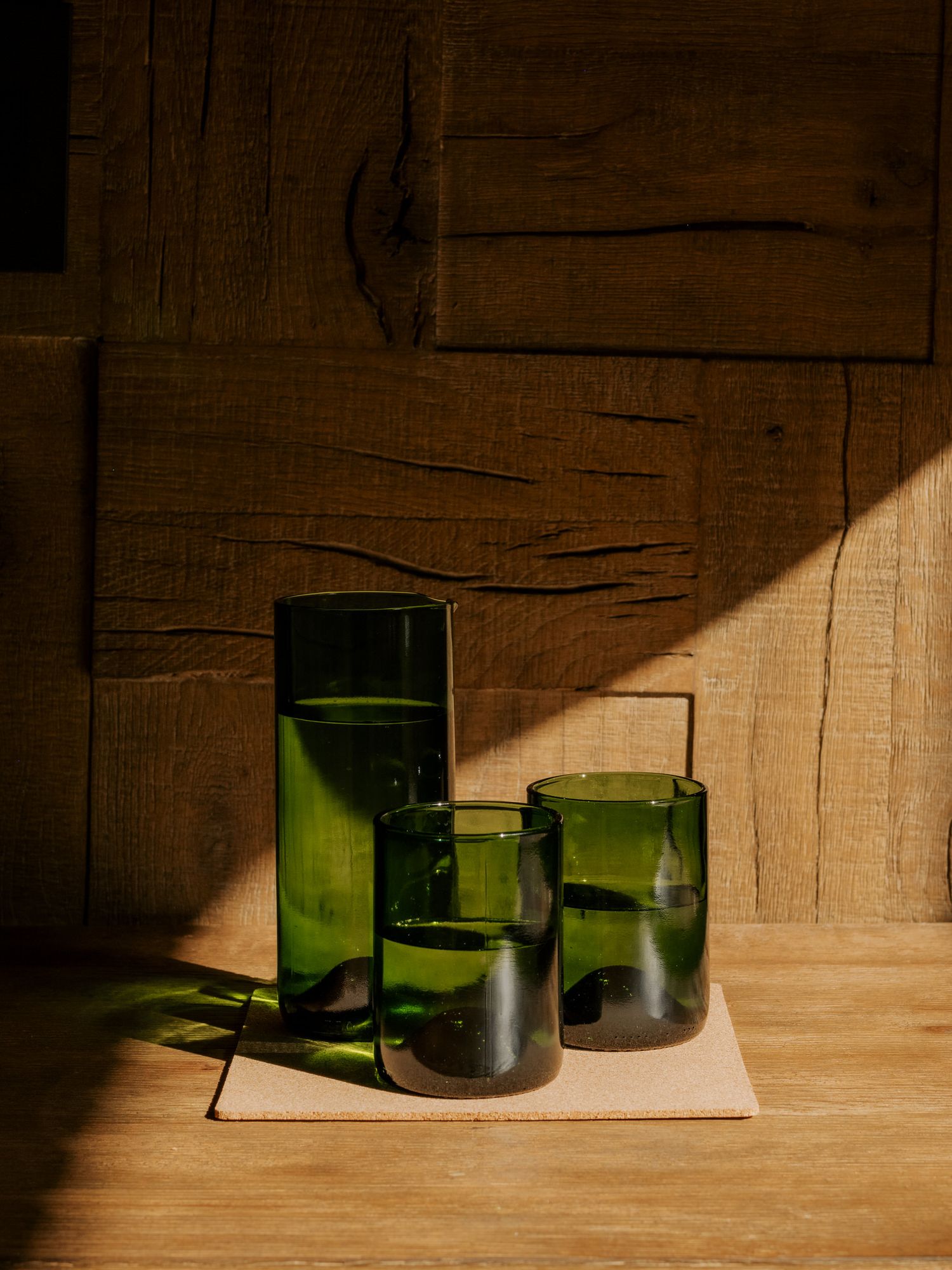 Three green glass tumblers of varying heights cast dramatic shadows on a wooden surface in moody lighting.