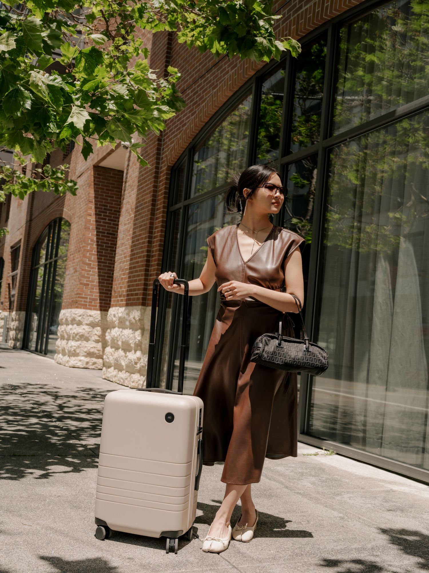 Urban traveler in brown dress stands beside modern white luggage on sunny city sidewalk near brick building.