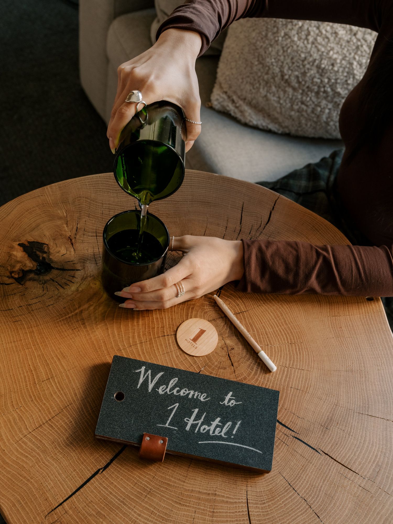Hands pouring wine at a wooden table with a chalkboard sign reading 'Welcome to I Hotel' near a grey pillow.