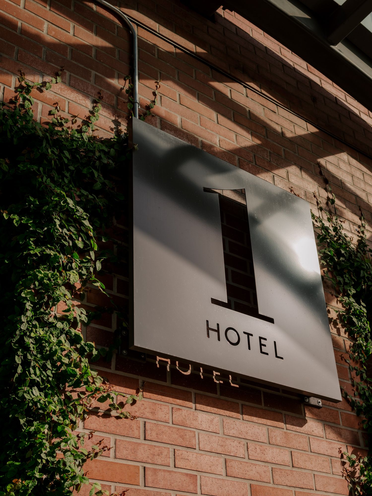 Modern minimalist hotel sign with black lettering mounted on white panel against exposed brick wall with climbing ivy.