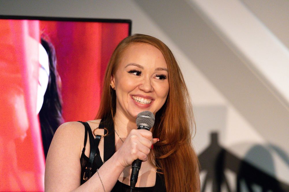 A person with long hair performs on stage with a microphone against a red-lit background.