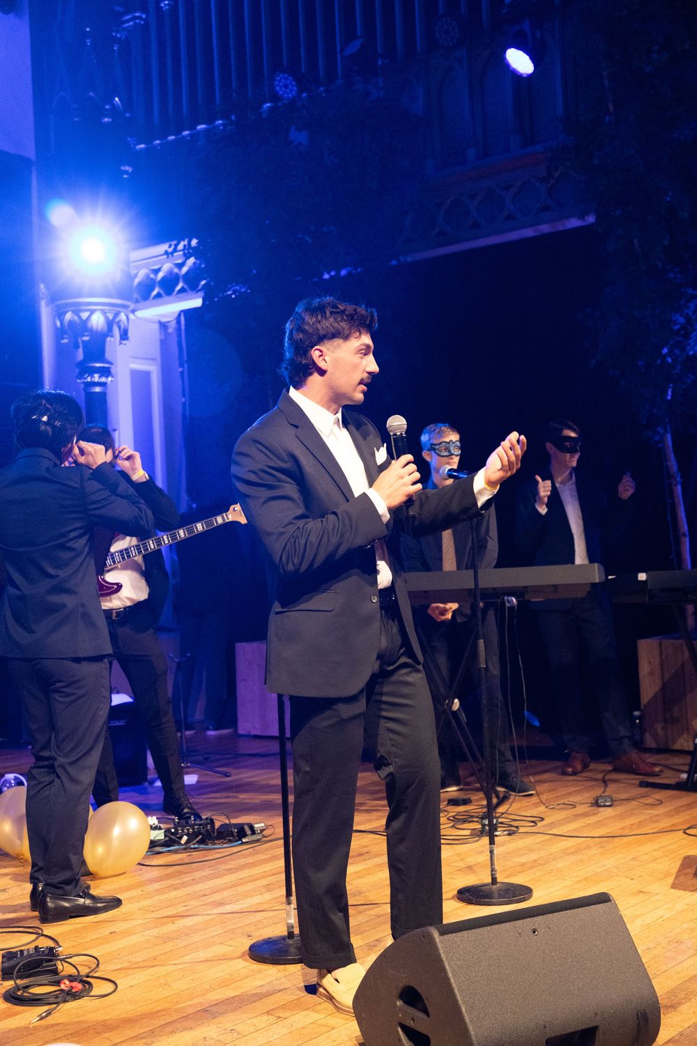 Performers in black suits singing on a wooden stage with blue stage lighting and microphone stands.