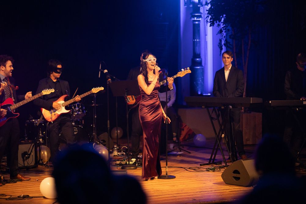 A performer in a burgundy dress sings on stage with a band during a nighttime concert performance with blue lighting.