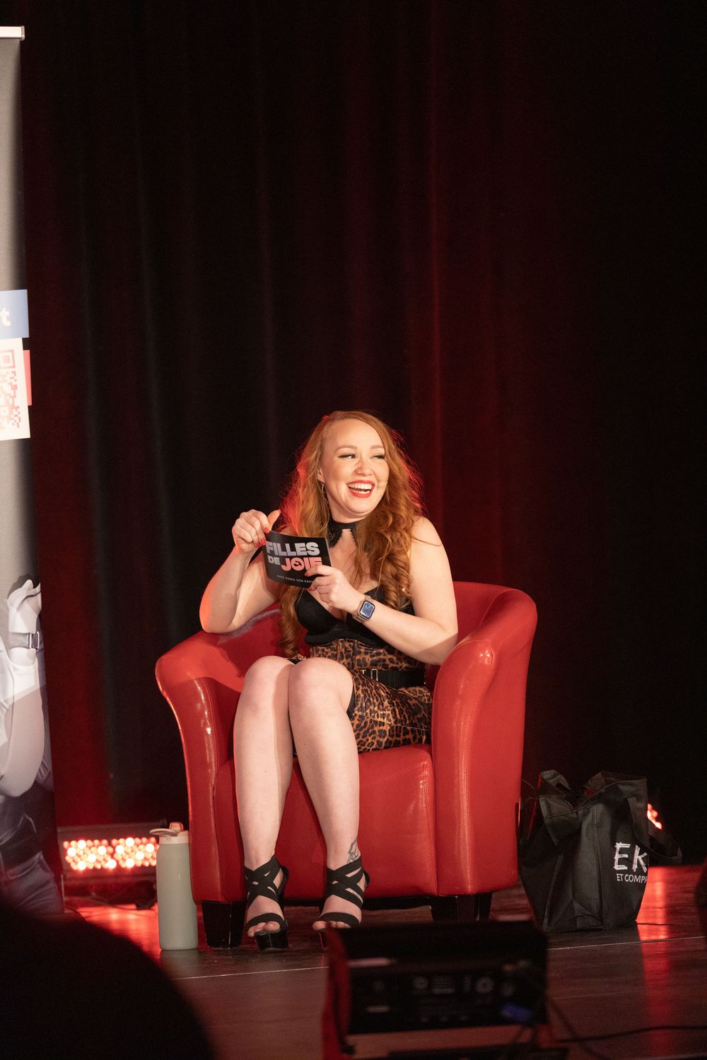 A person sits in a red armchair on a dark stage, appearing to be engaged in a lively conversation or performance.