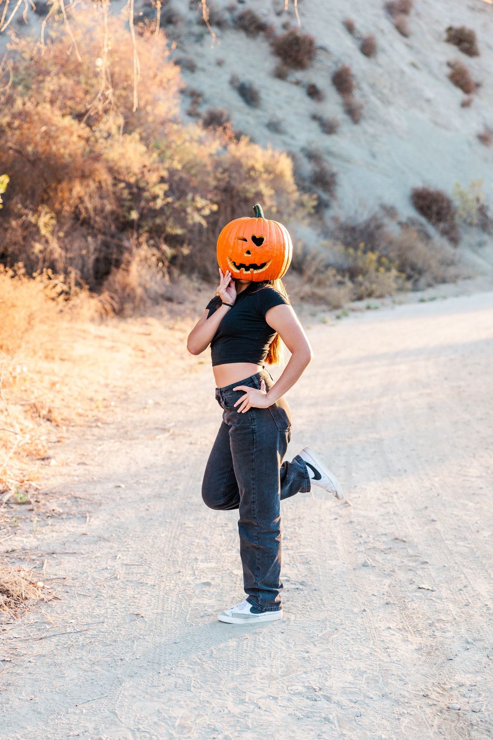 A person wearing a jack-o-lantern head and black clothes strikes a playful pose on a dirt path in autumn.