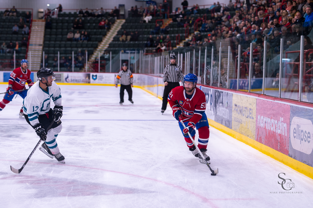 Hockey players from opposing teams chase the puck along the boards during intense gameplay.