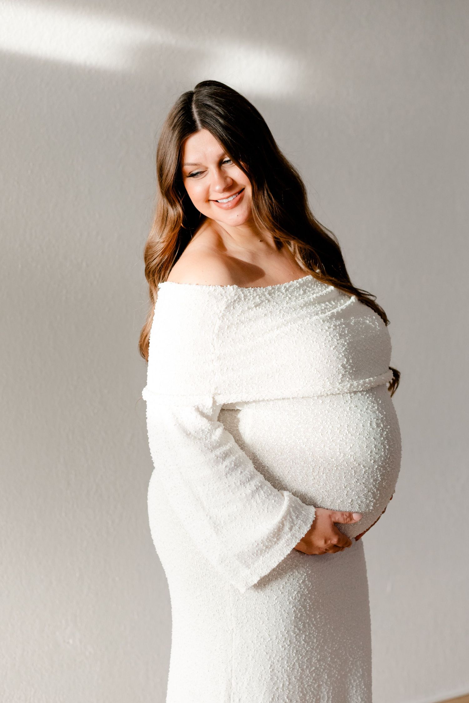 Smiling pregnant woman in white off-shoulder dress cradling baby bump in soft natural light.
