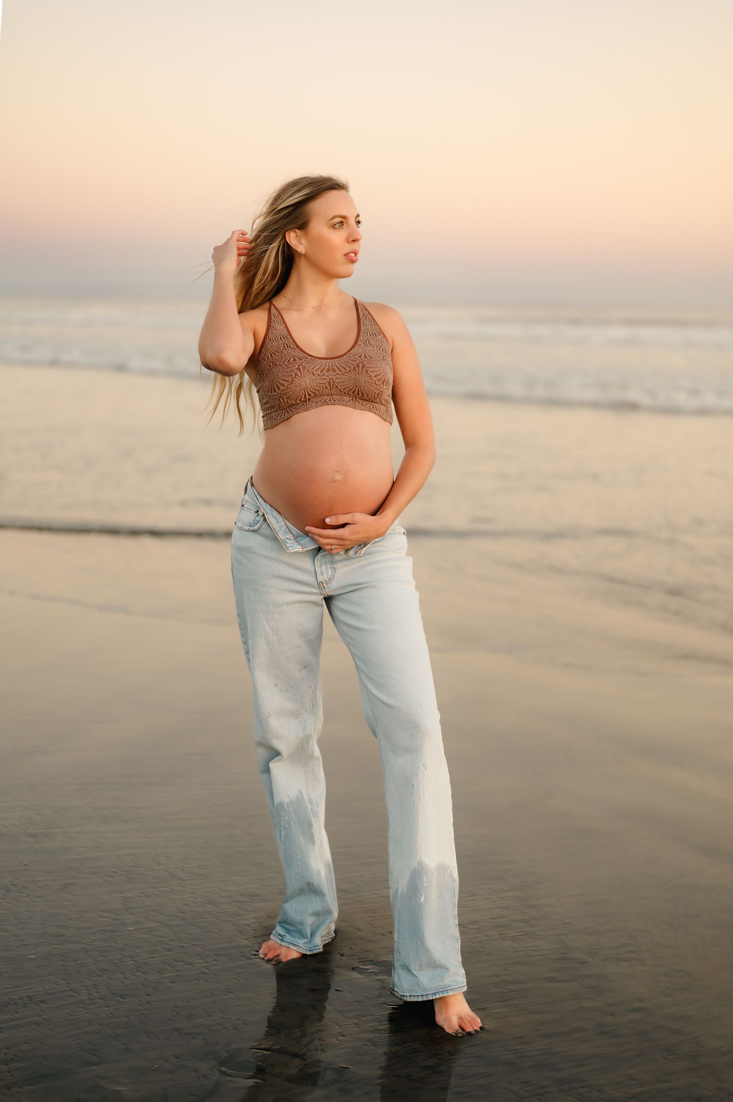 blue hour maternity photoshoot by Santa Barbara maternity photographer of woman in jeans and sports bra holding bare belly at Ventura beach