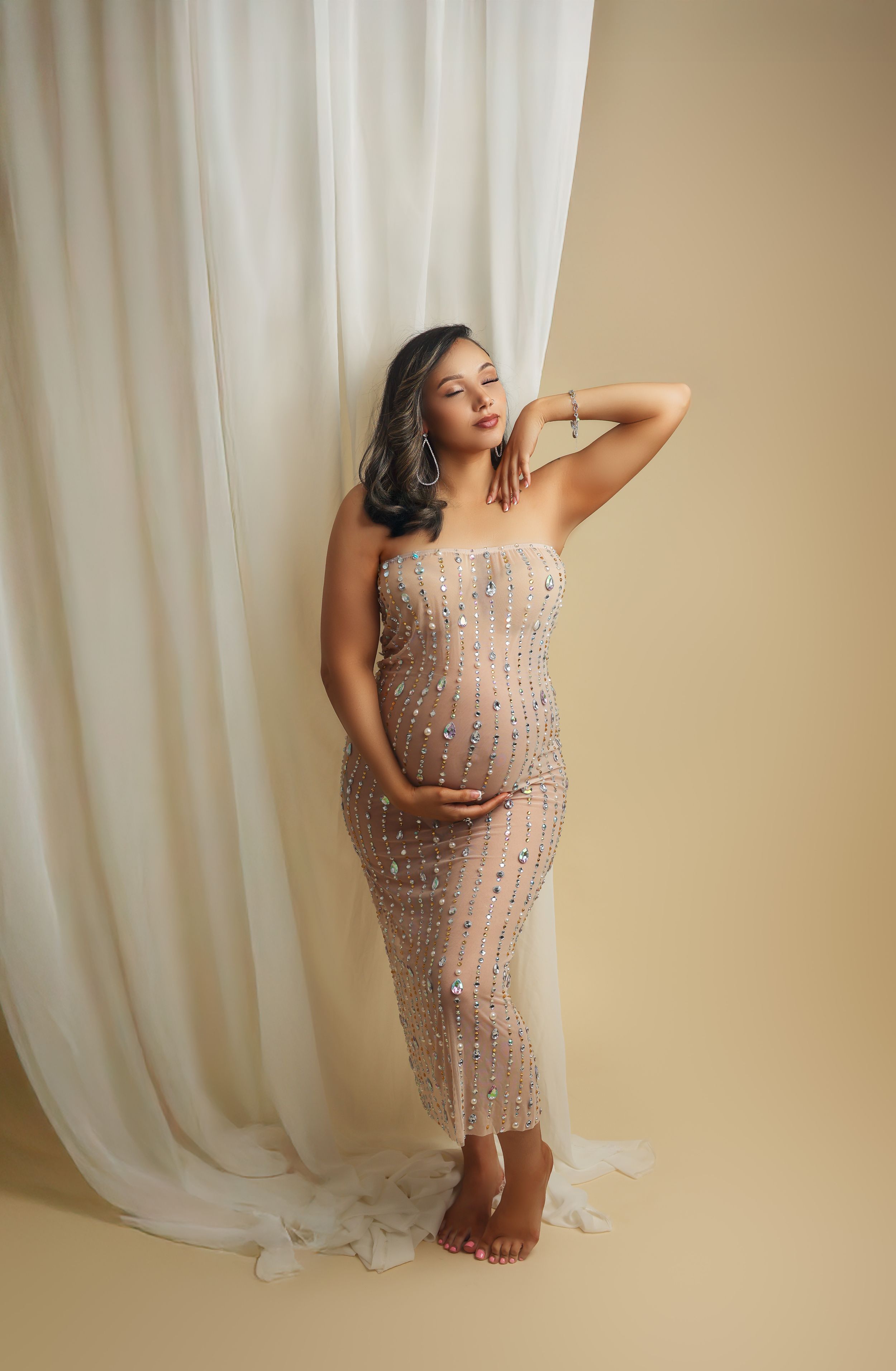 Expectant woman in sheer beaded gown poses gracefully against cream curtain backdrop during maternity photoshoot.