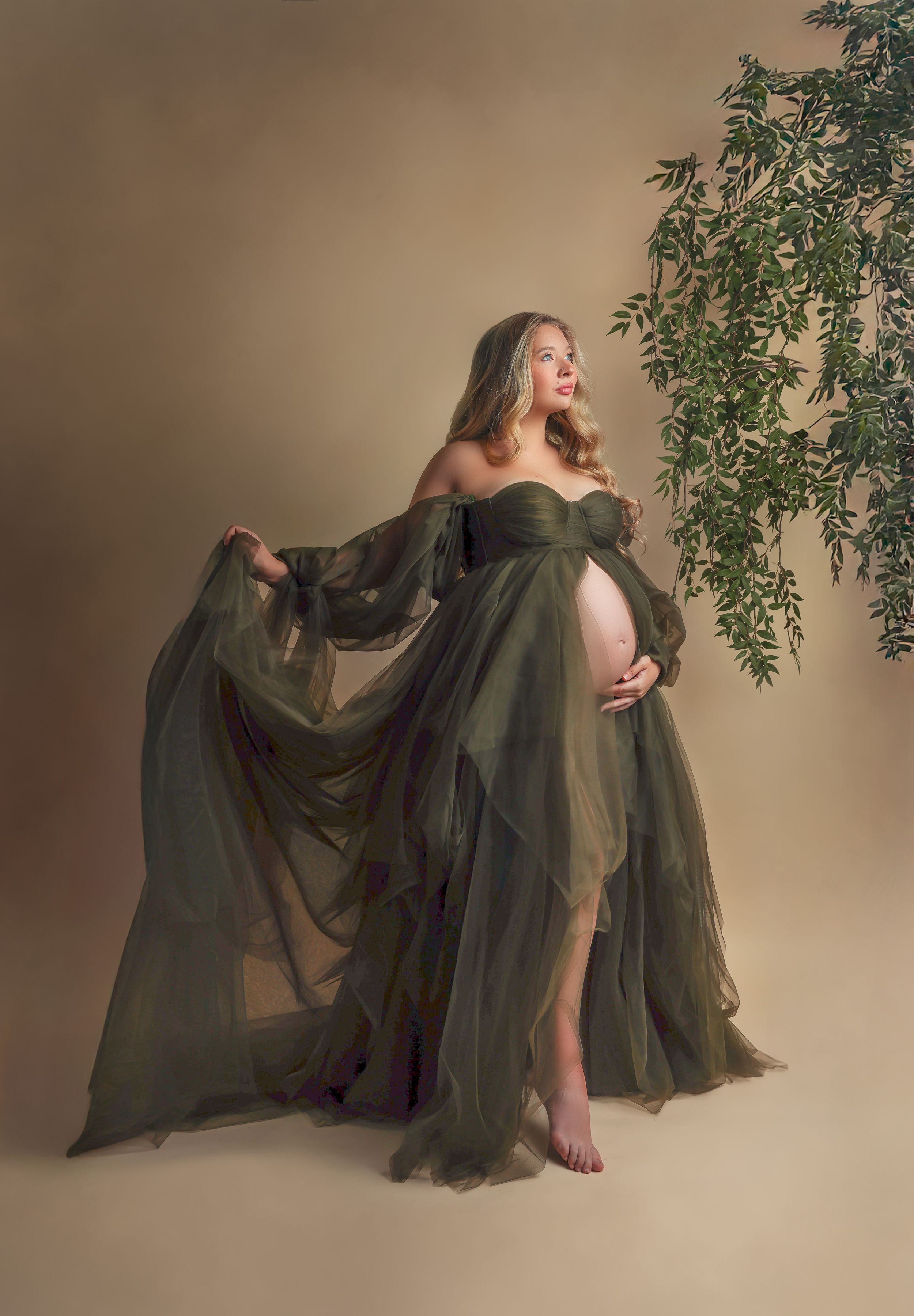 A flowing olive green gown creates an ethereal maternity portrait against a neutral studio backdrop with delicate foliage.