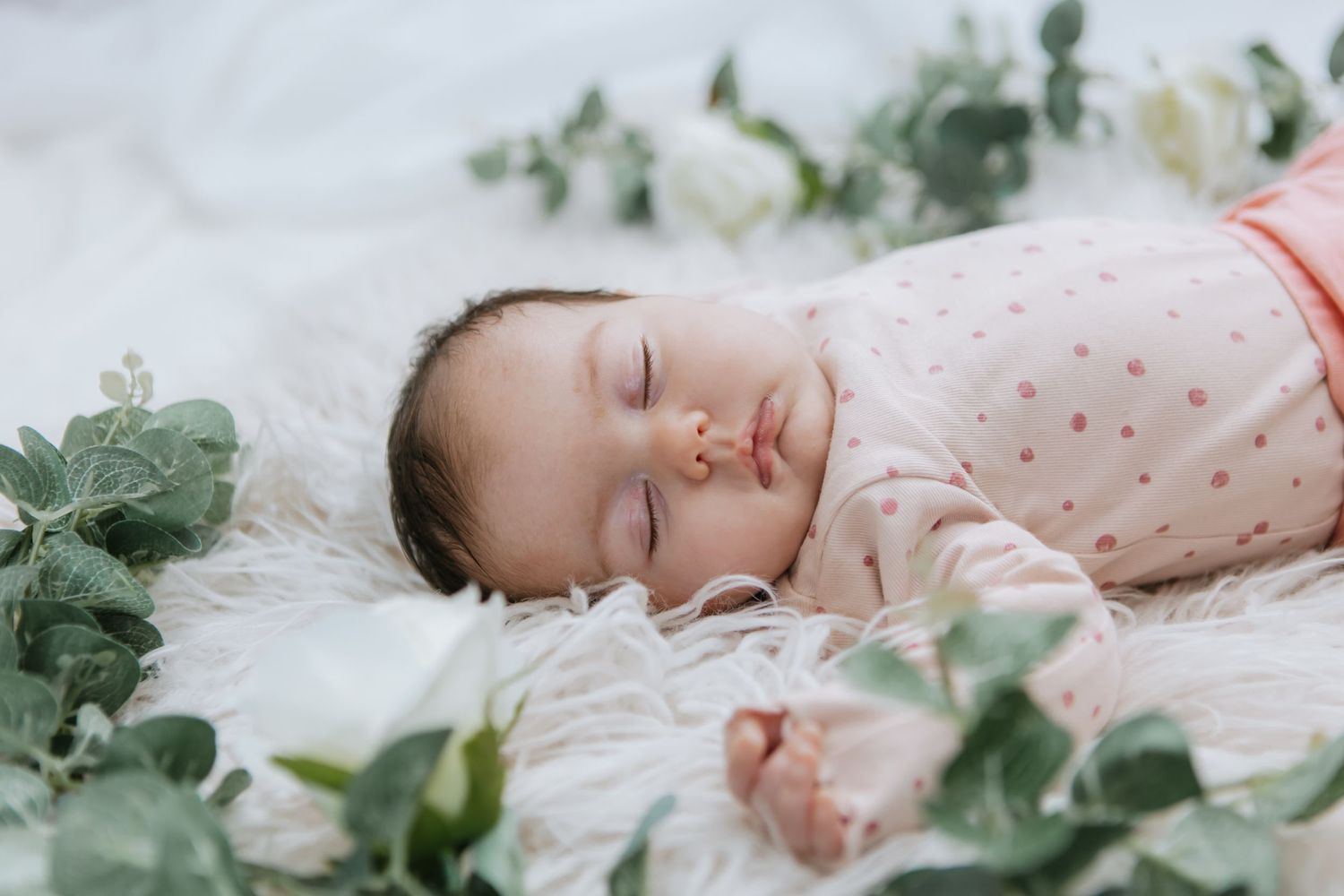 A baby sleeps peacefully on white bedding surrounded by green eucalyptus leaves and white flowers.