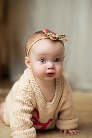 Baby wearing cream knit sweater and floral headband crawls on wooden floor in soft natural light.
