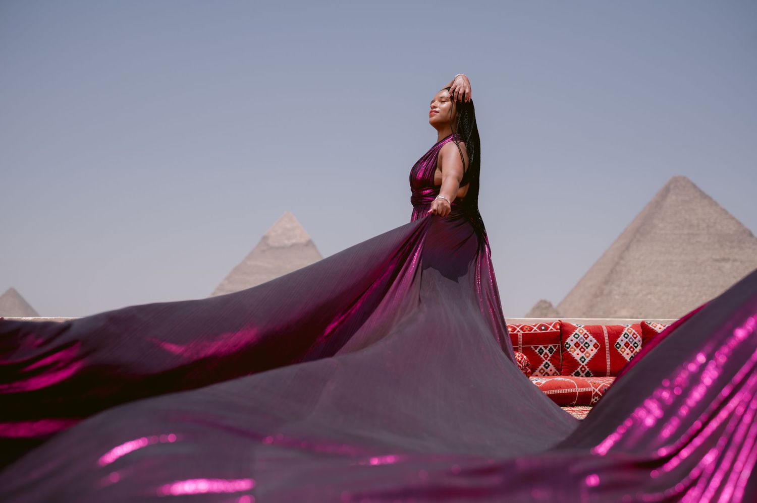 Magenta dress flows dramatically against sunset sky with pyramids silhouetted in background.