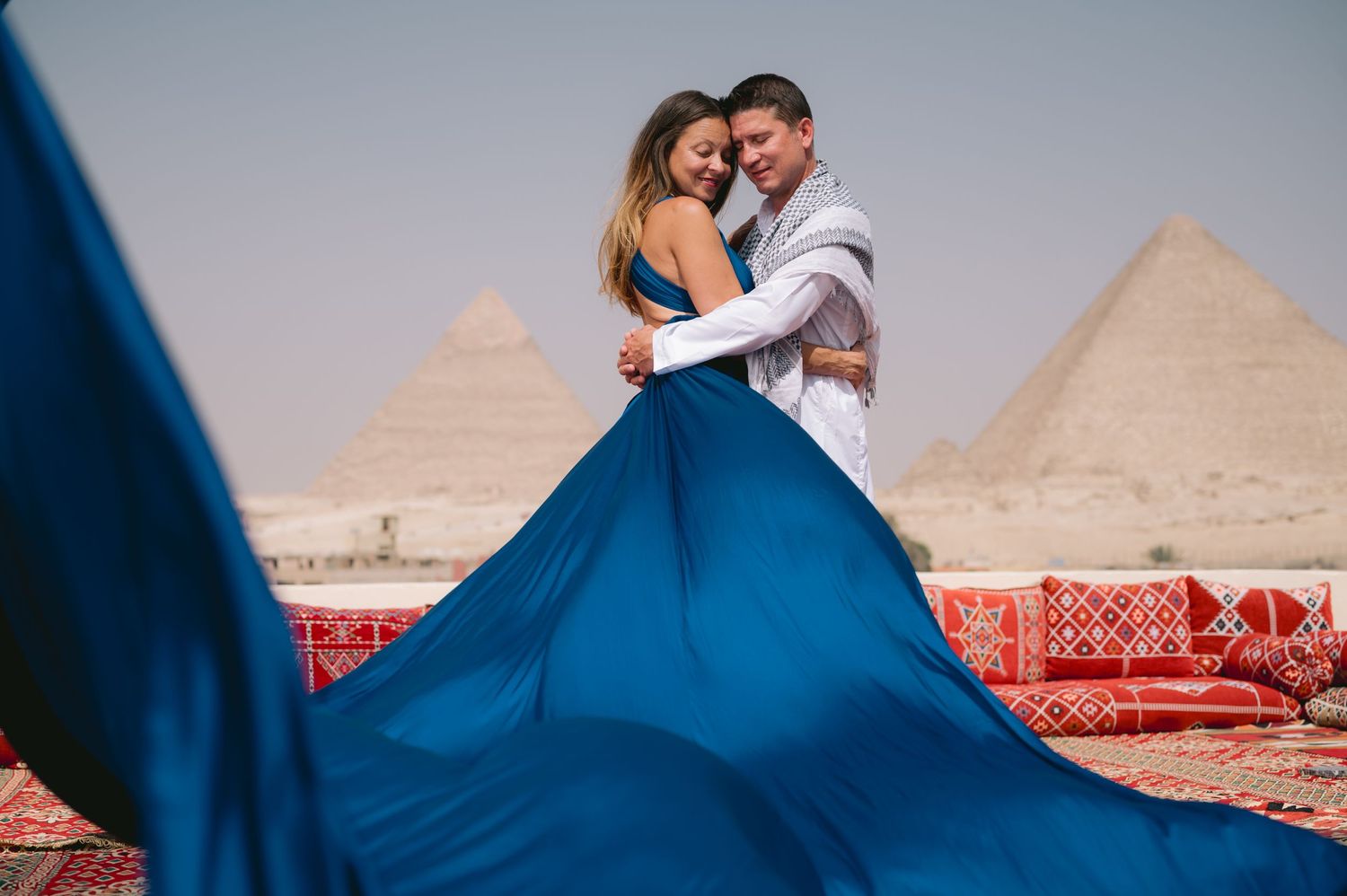 Royal blue dress billows dramatically as couple embraces with pyramids in background.