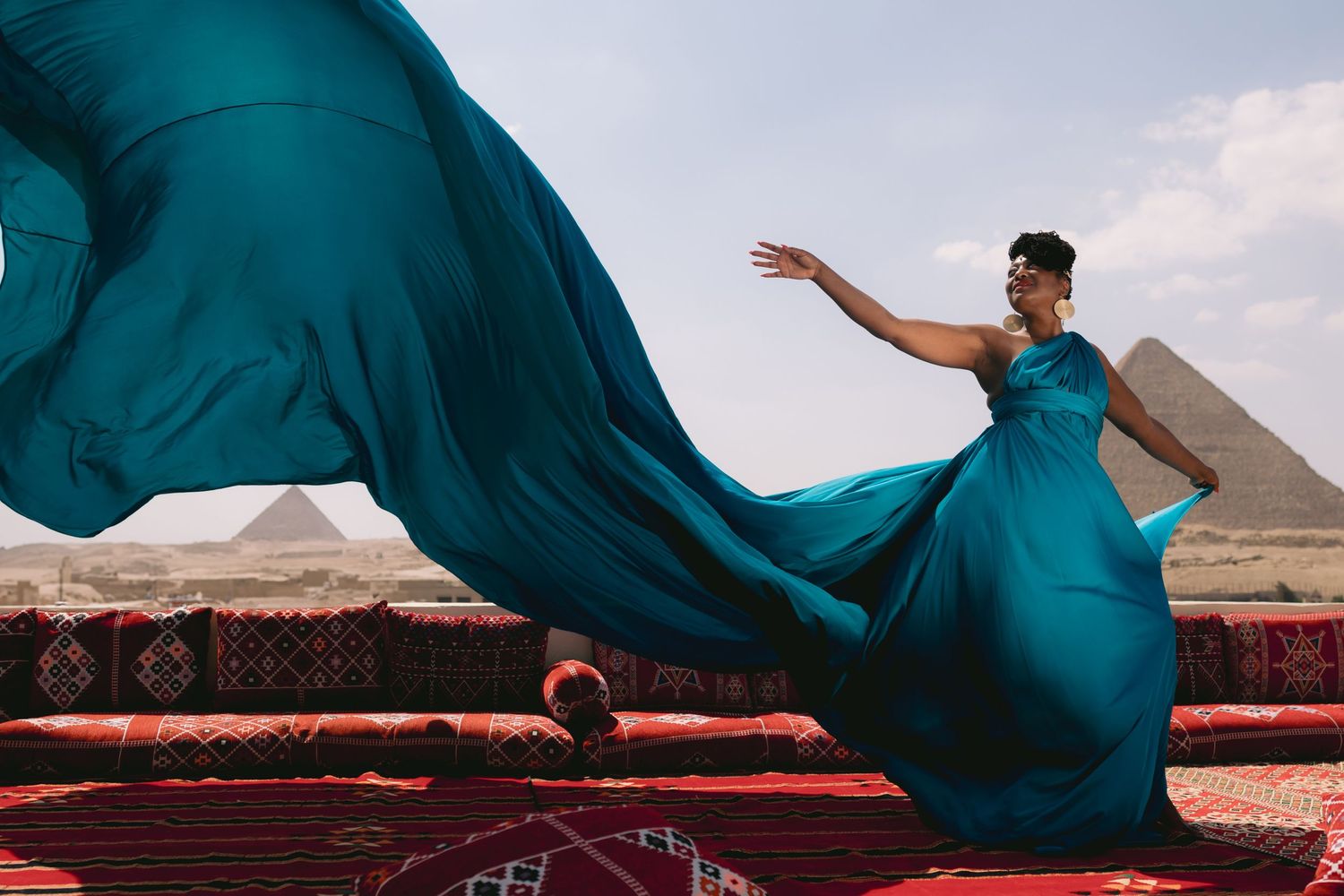 Teal dress flows in wind as figure poses on red carpet with pyramids in background.