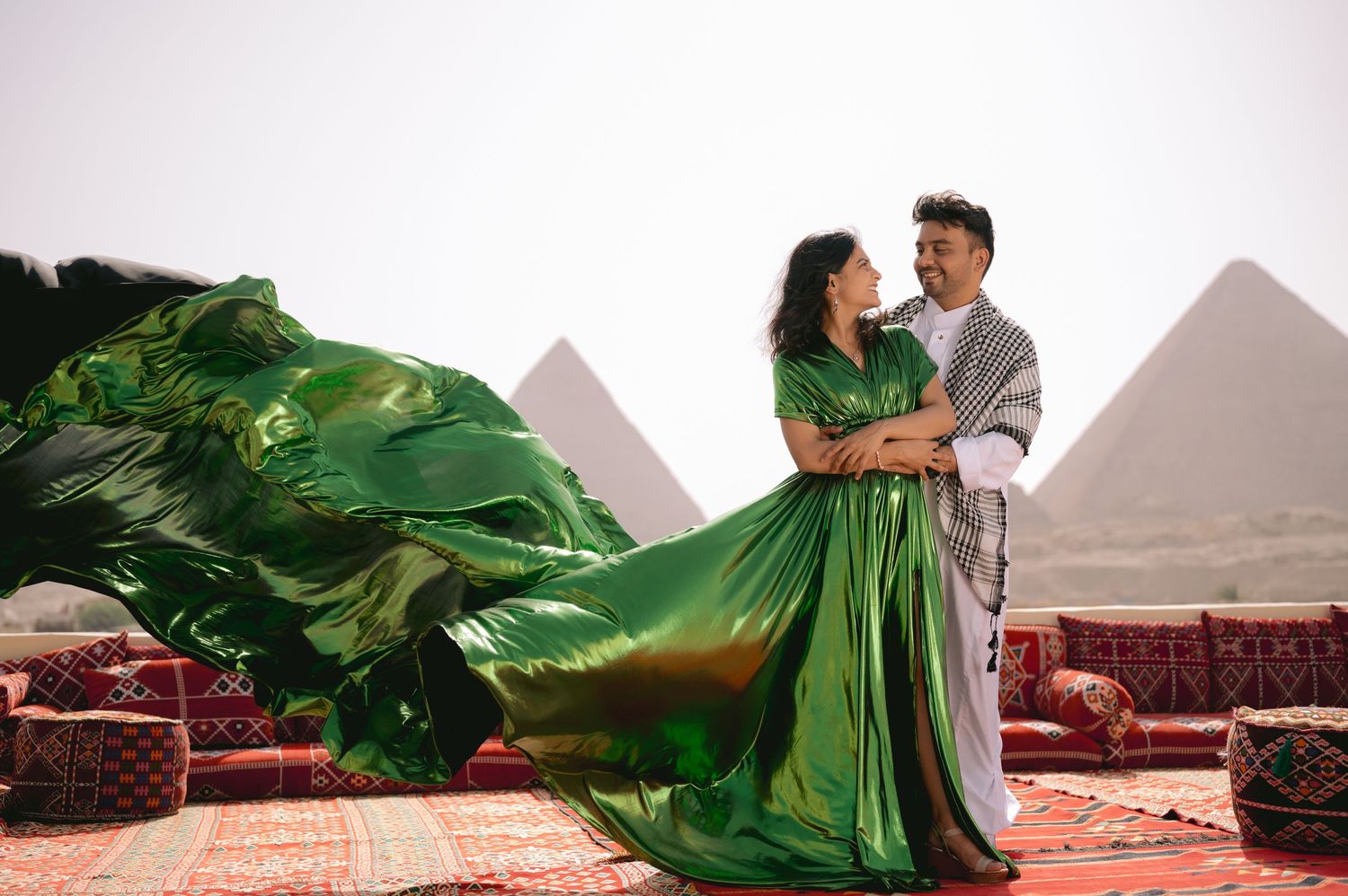 Flowing green dress catches wind as couple embraces with pyramids of Giza in background.