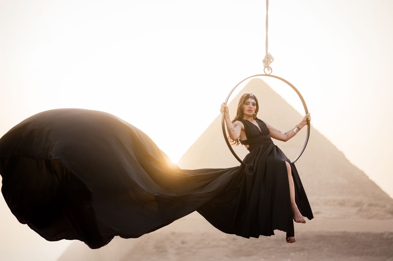 Flowing black dress billows dramatically as figure poses on aerial hoop with pyramid silhouette.