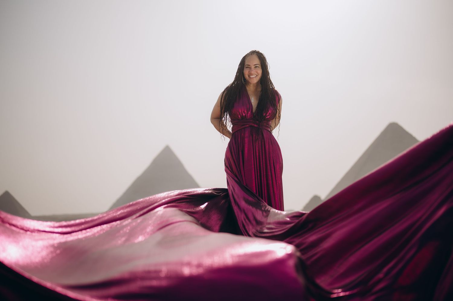 Woman in flowing burgundy gown poses dramatically against white backdrop with pyramids visible.