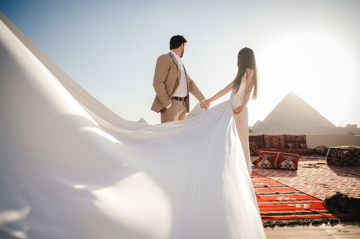 A couple in wedding attire poses with flowing dress fabric against the pyramids of Giza at sunset.
