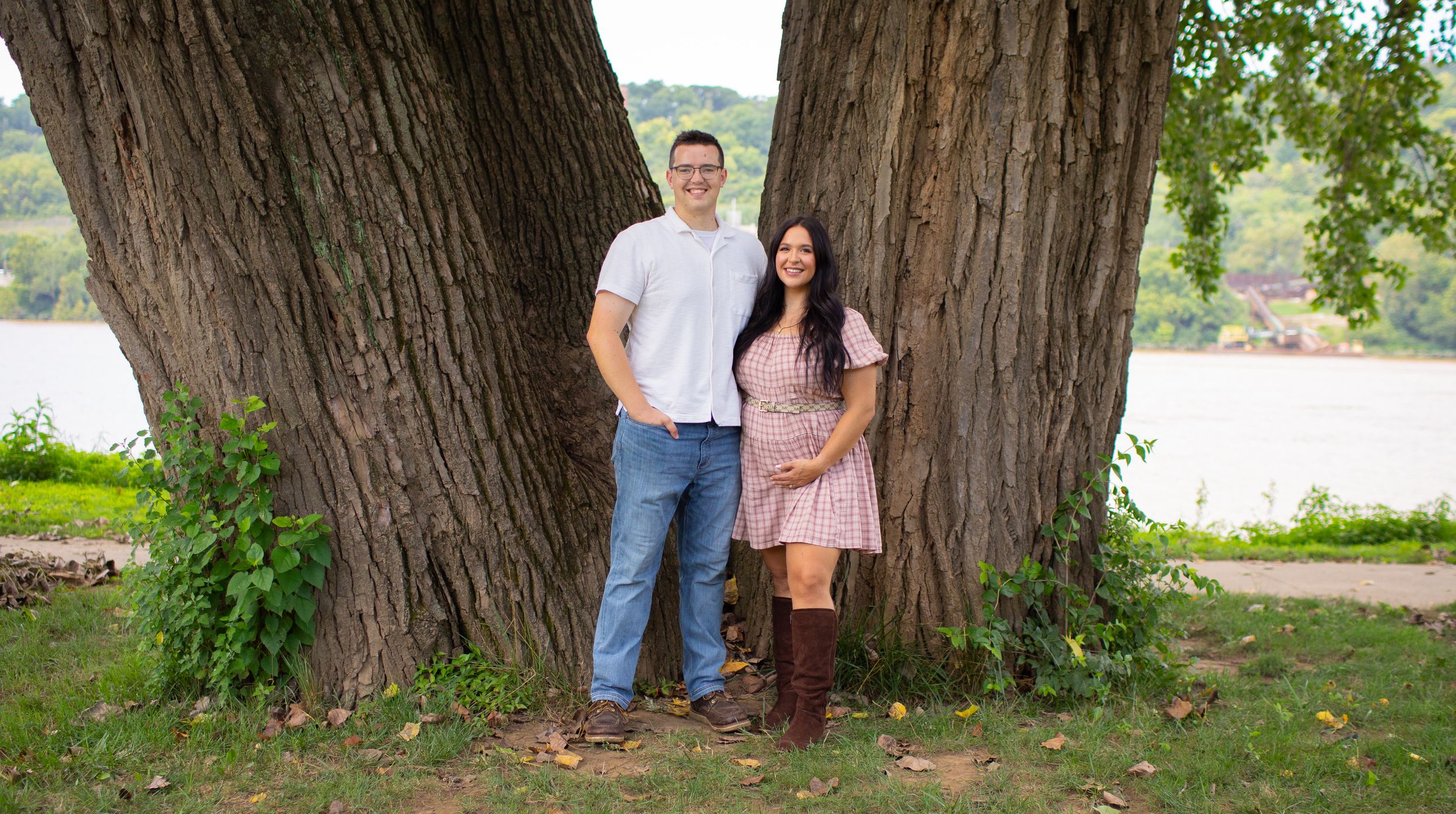 Couple stands together near large tree by a lake, woman in pink plaid maternity dress.