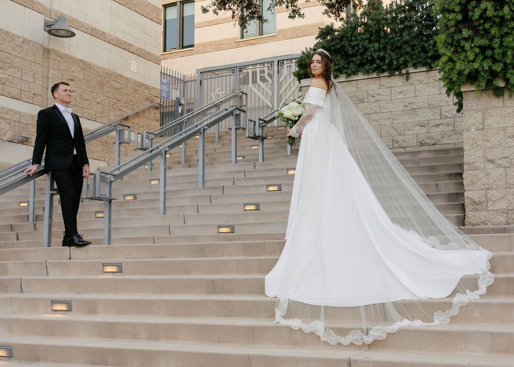 A bride in a flowing white gown walks gracefully down concrete steps at a stone building venue.