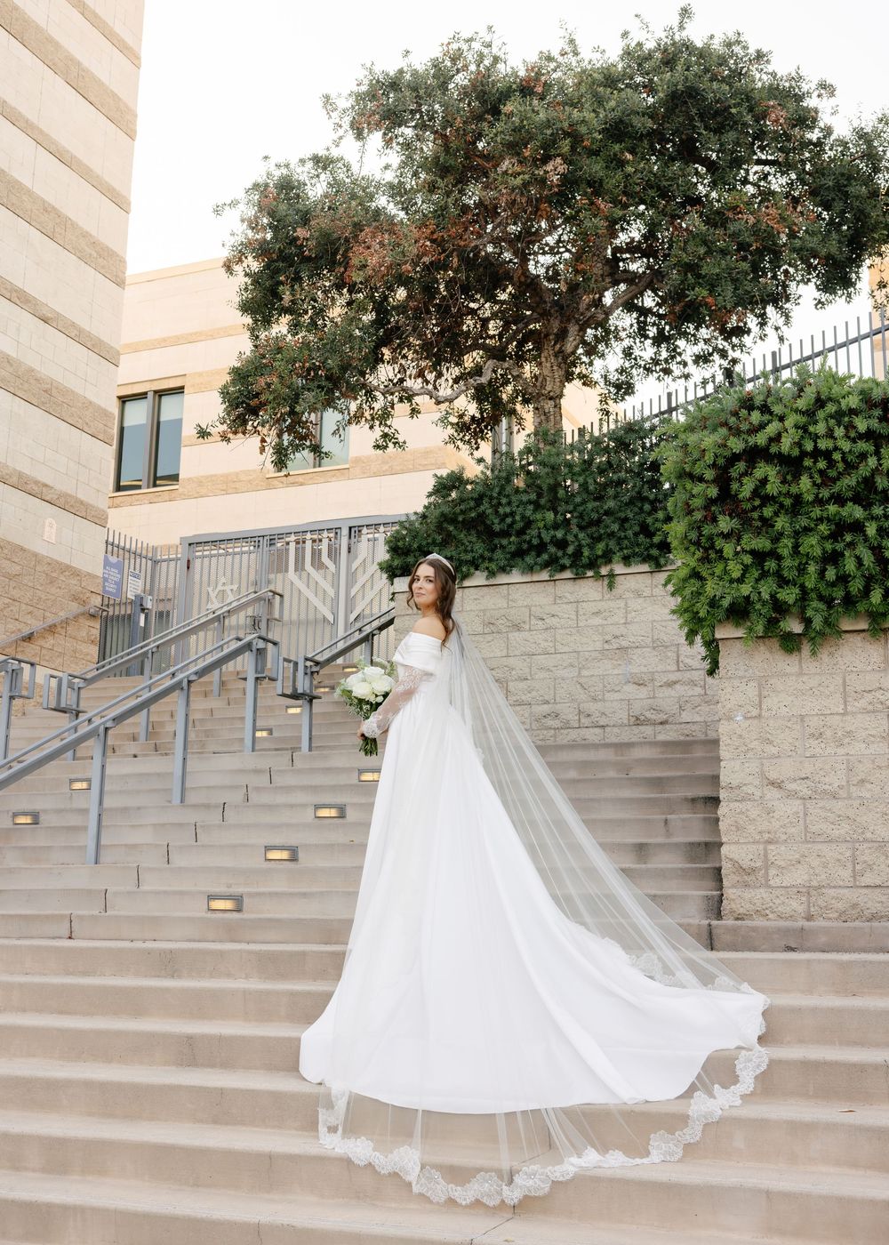 A flowing white wedding dress train cascades elegantly down stone steps against a garden backdrop.