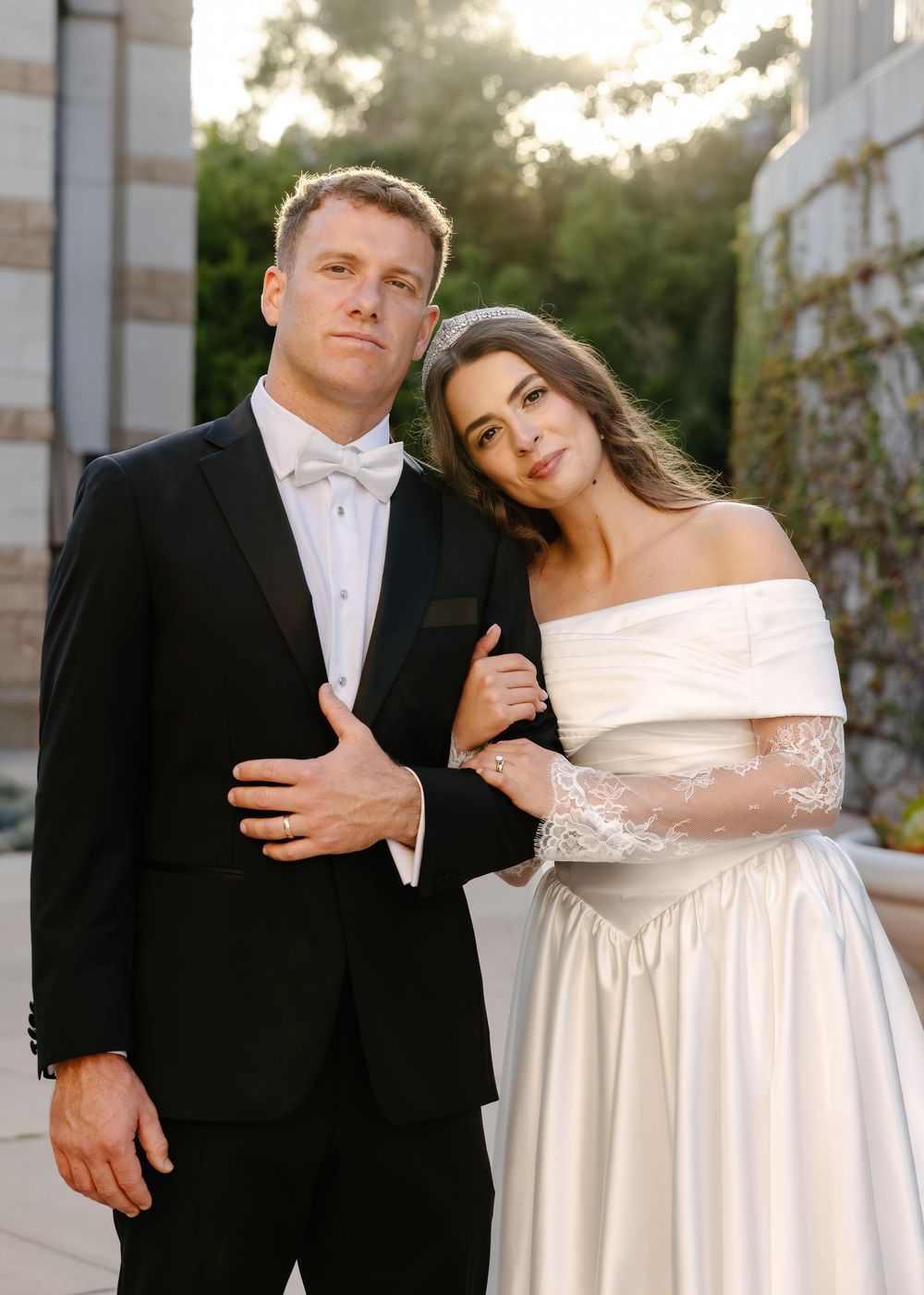 A couple in formal wedding attire leans against white pillars during a sunset photo shoot.