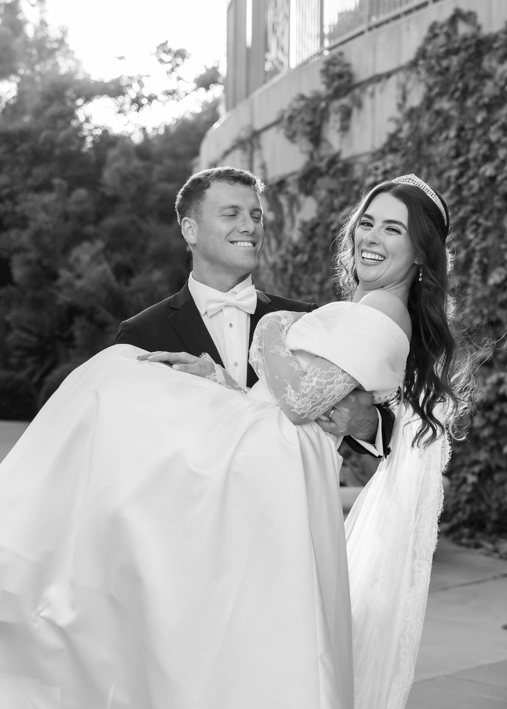 A black and white wedding photo captures a joyful moment with the groom carrying his bride in a flowing white dress.