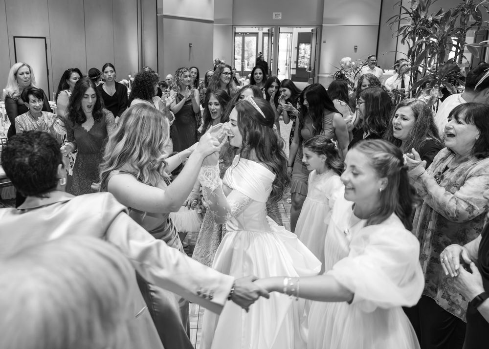 A large group of people in white clothes dance joyfully in an indoor celebration gathering.