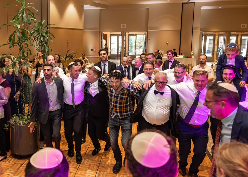 A group of wedding guests dance together under purple lighting at an indoor reception venue.