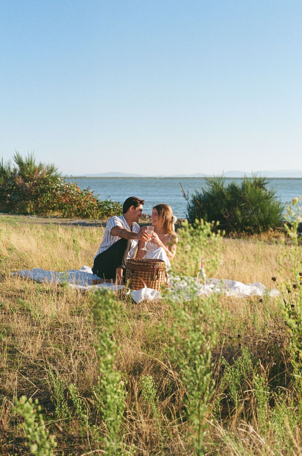 Couple sharing drinks during seaside picnic with scenic coastal view.