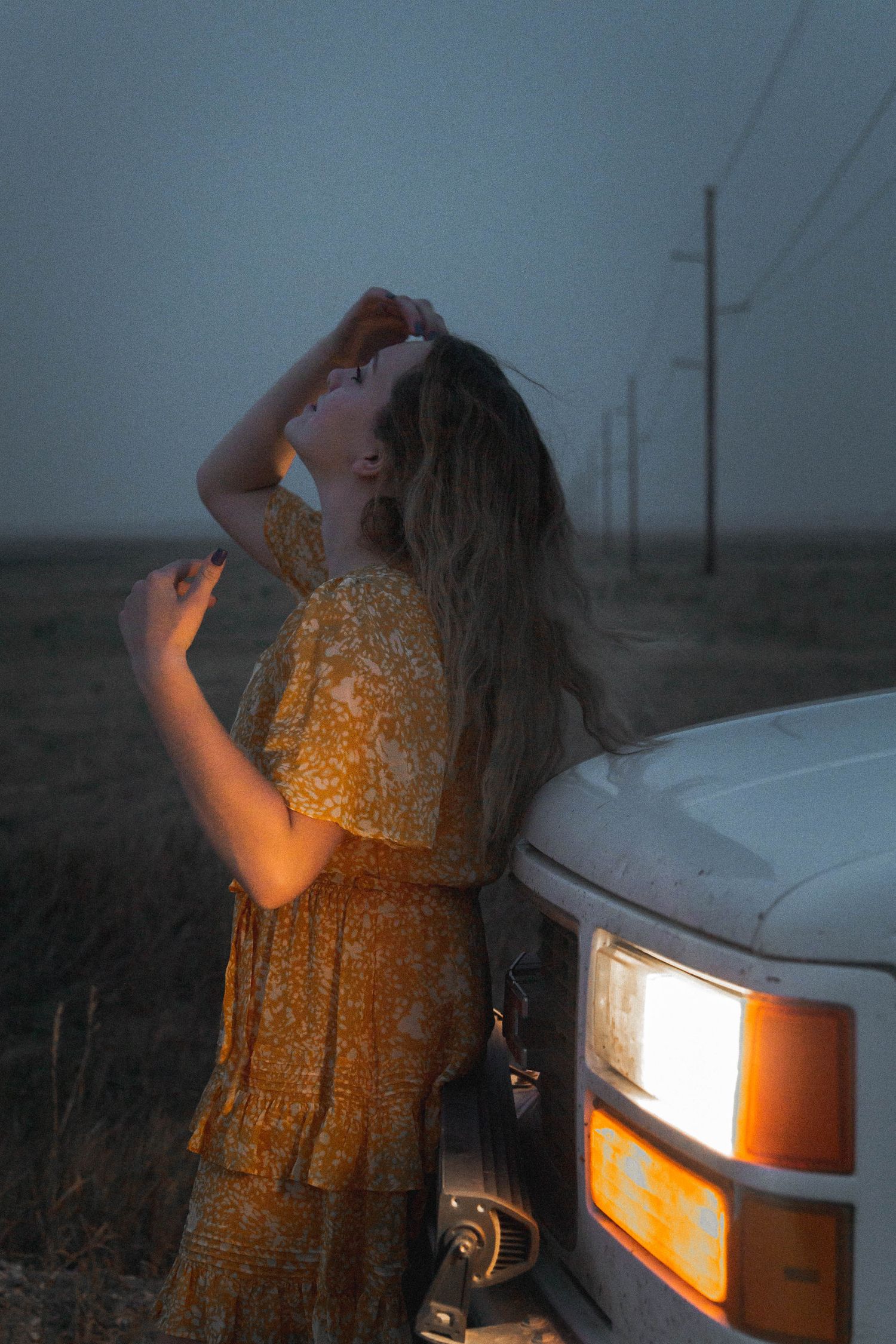 Young woman in a yellow floral dress leans against a white vehicle at dusk in a rural field setting.