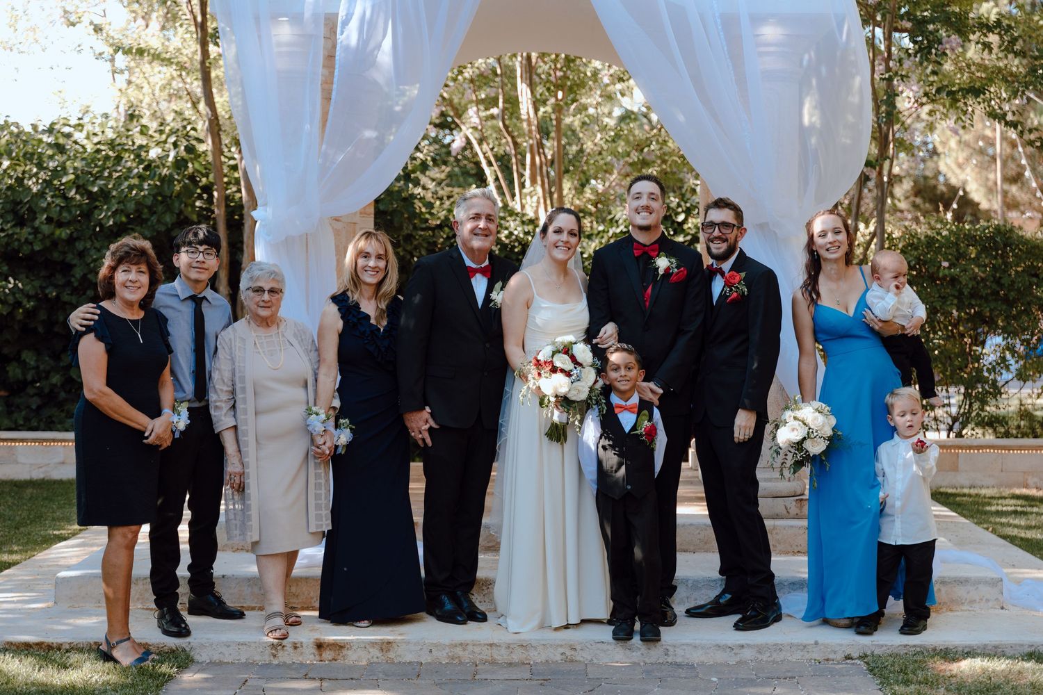 A formal wedding party portrait under a white arch in an outdoor garden setting.
